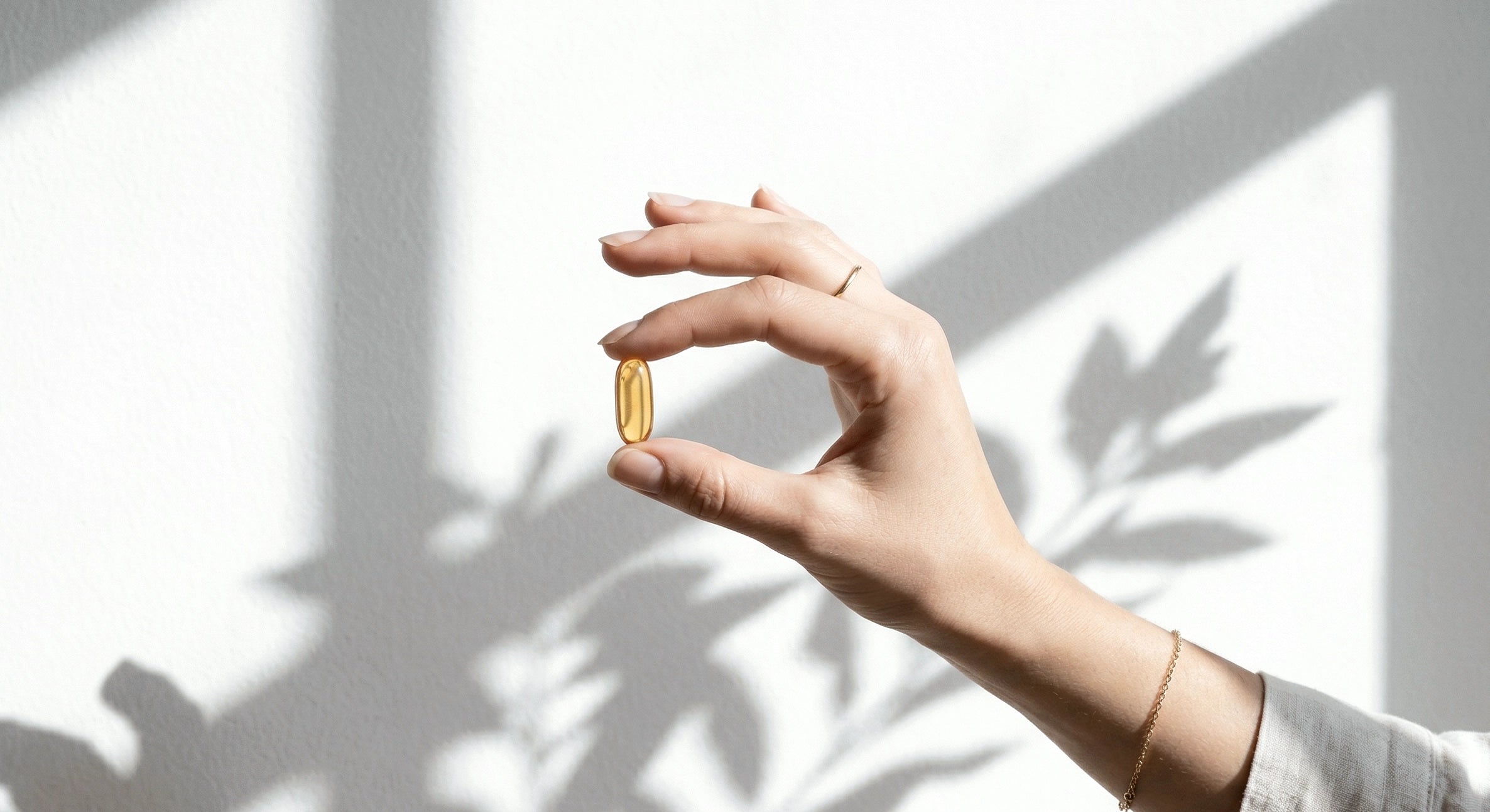 Genviten norvegiškų žuvų taukų kapsulė rankoje šešėlių fone – skaidri, aukštos koncentracijos Omega-3 trigliceridų forma be jokių dirbtinių priedų.